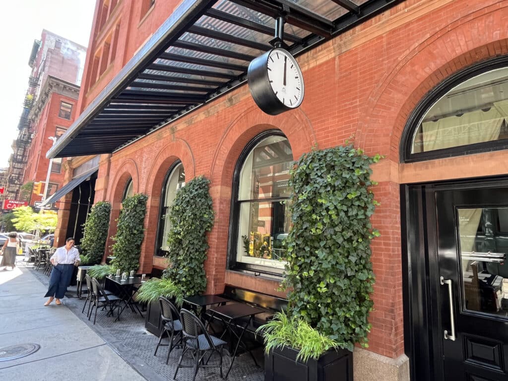 NYC NY- double sided bracket clock hanging from entry way covering with tables and chairs and ivy growing on the exterior of a brick wall with person smiling