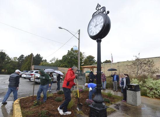 People with shovels planting around the base of a tower clock, near a parking lot and brick wall