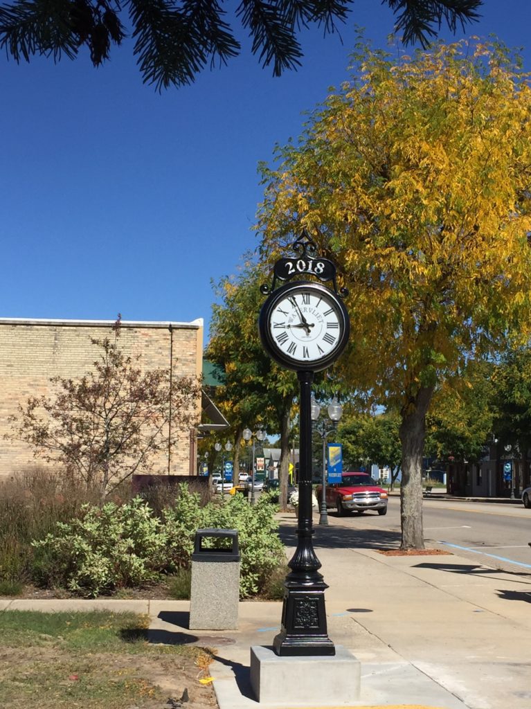 A  Lumichron Post Clock for the town of Watervliet, Michigan. Victorian style double-faced streetscape clock with custom name panel, shown installed on street with blue sky, trees, and building in the background