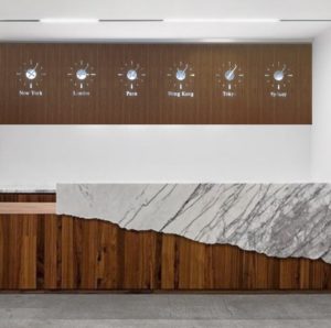 A marble and wood desk with a background of wood paneling, with set of 6 stainless steel wall clocks, each set to a world time New York, London, Paris, Hong Kong, Tokyo, and Sydney