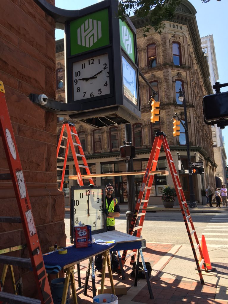 Vintage McClintock Clock repair in process, on a street corner with clock repairman and ladders. 4-way clock has 30-inch square faces, one removed for cleaning.