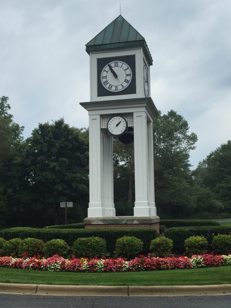 This is a 4-way clock tower at the entrance to Wildwood Springs community. A white frame tower with a brick base and topped with a green roof, surrounded by a garden, the tower has 4 clocks and a thermometer