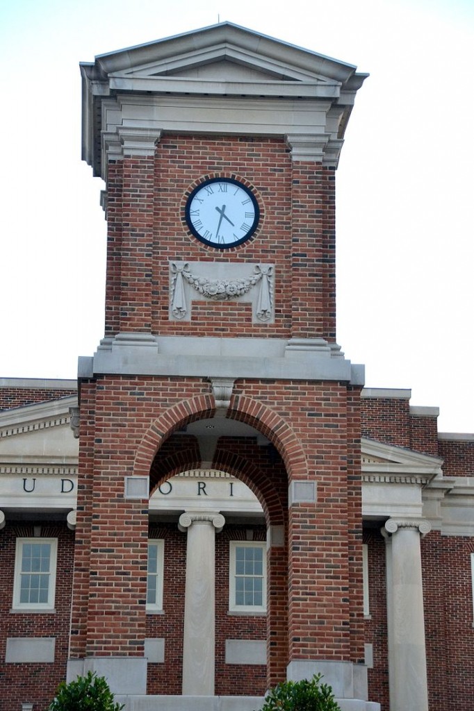 A neo-classical style brick clock tower with tower clock in front of a brick auditorium at the University of Alabama